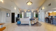 This single-floor community clubhouse offers a well-appointed layout across seven distinct areas, designed to serve as a functional and inviting gathering space. The main lounge is a standout feature, boasting soaring vaulted ceilings, a ceiling fan with a central pendant light, and soft blue-gray walls that create a relaxed coastal ambiance. Wicker furniture with navy blue cushions and patterned throw pillows adds a warm, casual elegance, while large windows dressed with decorative floral curtains flood the space with natural light and offer views to the outdoors. Large-format tile flooring flows throughout, lending a clean, cohesive look. A secondary seating area features bold red leather sofas, a modern round pedestal table, and floor-to-ceiling windows with French doors that open to the exterior, bathing the room in light. The kitchen is equipped with granite countertops, a mosaic tile backsplash, warm wood cabinetry, and full-size black appliances, including a refrigerator and dishwasher. A dedicated business center offers a row of computer workstations along a built-in desk beneath a window, accented by coastal-themed artwork. Glass-paned French doors connect several zones, enhancing the open, airy feel throughout this thoughtfully designed community facility.