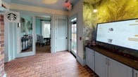 This inviting cyber cafe exudes a warm, eclectic character through a thoughtful blend of classic architectural elements and comfortable furnishings. Throughout the space, rich terracotta brick flooring sets a rustic, welcoming tone that ties the entire area together seamlessly.

A standout feature is the striking brick archway that frames the transition between zones, adding a timeless, artisanal quality to the interior. Crisp white columns with pale aqua-tinted crown molding and baseboards add a refined Southern charm, complementing the soft, light-toned walls.

The lounge area offers a relaxed atmosphere with vintage-style leather wingback chairs arranged around a dark wood side table, all positioned beneath a classic pendant light and in front of large windows dressed with patterned drapery panels and wooden blinds that invite generous natural light.

A bold tropical botanical wallpaper energizes one wall near the service counter, where a dark granite-topped station with soft gray cabinetry provides a functional check-in or ordering area with a digital display. A brick-framed niche houses a coffee station complete with a beverage cooler underneath, adding convenience and character.

Ceiling fans contribute to the relaxed ambiance, while glass-paneled doors and windows throughout maintain a bright, airy feel and offer views to the exterior, completing this charming and well-appointed space.