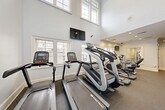 Located at 100 Cavalier Crossing in Stonecrest, GA, this fitness center offers a thoughtfully designed exercise space across a single floor. The room features soaring double-height ceilings in the cardio area, creating an open and airy atmosphere enhanced by large windows that flood the space with natural light and offer views of the surrounding exterior. The flooring throughout is durable rubber gym matting, practical for both cardio and strength training activities. Recessed lighting and ceiling fans ensure the space remains well-lit and comfortable during workouts. The cardio section is equipped with multiple treadmills, stair climbers, elliptical trainers, and stationary bikes, including Star Trac branded machines, accompanied by wall-mounted flat-screen televisions for entertainment. The strength training area is lined with floor-to-ceiling mirrors that visually expand the space, and features a comprehensive array of cable and weight machines. Decorative diagonal stripe accents on one wall add a modern, energetic touch to the overall aesthetic. Crown molding and clean white walls give the space a polished, upscale feel. A hand sanitizer dispenser and posted facility rules reflect attention to cleanliness and user safety. The fitness center provides a complete workout environment suited for a wide range of exercise routines.