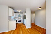 Located at 13836 NE 8th St in Bellevue, WA, this single-floor, one-bedroom unit offers a well-designed layout with consistent warm hardwood flooring flowing throughout every room. The kitchen is configured as a galley-style space, outfitted with crisp white cabinetry featuring brushed nickel hardware, light quartz countertops, stainless steel appliances including a refrigerator, range, microwave, and dishwasher, and a window above the sink that brings in natural light. The adjoining living room is a comfortable and inviting space anchored by a classic fireplace with a white mantle and dark stone surround. Windows facing the outdoors allow natural light to fill the room, enhancing its bright and airy feel. A hallway connects the main living areas to the generously sized primary bedroom, which offers ample square footage, neutral-toned walls, and windows that provide views of the surrounding neighborhood. The overall aesthetic of the unit is clean and modern, with a neutral color palette of whites and soft grays that creates a cohesive, move-in-ready atmosphere throughout the space.