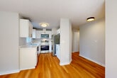 Located at 13836 NE 8th St in Bellevue, WA, this single-floor, one-bedroom unit offers a well-designed layout with consistent warm hardwood flooring flowing throughout every room. The kitchen is configured as a galley-style space, outfitted with crisp white cabinetry featuring brushed nickel hardware, light quartz countertops, stainless steel appliances including a refrigerator, range, microwave, and dishwasher, and a window above the sink that brings in natural light. The adjoining living room is a comfortable and inviting space anchored by a classic fireplace with a white mantle and dark stone surround. Windows facing the outdoors allow natural light to fill the room, enhancing its bright and airy feel. A hallway connects the main living areas to the generously sized primary bedroom, which offers ample square footage, neutral-toned walls, and windows that provide views of the surrounding neighborhood. The overall aesthetic of the unit is clean and modern, with a neutral color palette of whites and soft grays that creates a cohesive, move-in-ready atmosphere throughout the space.