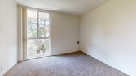 This single-floor residence offers a thoughtfully organized layout that balances comfort and functionality. The living area is generously sized, finished with neutral wall-to-wall carpeting that adds warmth underfoot, and features a wall-mounted heating unit alongside a sliding glass door that opens to a private balcony — providing pleasant views of surrounding trees and an abundance of natural light.

The kitchen is neatly tucked into a defined alcove, separated from the living space by a partial wall. It is equipped with white cabinetry, a white electric range, and light-toned countertops, all set atop tile flooring that transitions smoothly into the carpeted living zone. A flush-mount ceiling light illuminates the area effectively.

A carpeted hallway connects the living spaces to the private quarters, where a bedroom features large windows that frame leafy, tree-lined views, filling the room with natural light. A wall-mounted vent ensures year-round comfort.

The bathroom area includes a vanity with warm wood cabinetry and a granite-style countertop, complemented by a large mirror. Adjacent to the vanity is a built-in shelving unit offering practical storage solutions. The overall color palette throughout the residence is light and neutral, creating a clean, open, and inviting atmosphere well-suited for comfortable everyday living.