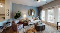Located at Eagle Hall Lane in Goose Creek, South Carolina, this single-story residence offers a thoughtfully designed layout visible across its interconnected living spaces. The living room serves as the centerpiece of the unit, featuring warm hardwood-style flooring, a ceiling fan, and French doors that open to a balcony, allowing natural light to fill the space. Neutral walls are accented by a soft blue feature wall, creating a calm, inviting atmosphere complemented by patterned drapery and a decorative area rug.

The kitchen is outfitted with modern gray flat-front cabinetry, granite-style countertops, a subway tile backsplash, and a full suite of stainless steel appliances, including a side-by-side refrigerator, range, and over-the-range microwave. The layout provides functional counter space and ample storage.

Two bedrooms each feature plush carpeting, large windows with blinds and coordinating curtains that bring in natural light and outdoor views, and sufficient space for full bedroom furnishings. The bathroom includes hardwood-style flooring, a granite-topped vanity with gray cabinetry, and a full tub-shower combination with a curved curtain rod. The overall aesthetic throughout is cohesive, modern, and residential.