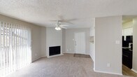 This single-floor residence offers a well-organized and functional layout throughout. The living area serves as the heart of the home, featuring a classic white brick fireplace that anchors the space with warmth and character. A ceiling fan with integrated lighting provides both comfort and illumination, while neutral-toned carpet and soft wall colors create a clean, welcoming atmosphere. A textured popcorn ceiling adds a traditional finish overhead. The entry door opens directly into this main living space, which connects seamlessly to the kitchen area via an open pass-through layout. The kitchen is equipped with dark appliances, including a refrigerator, and is framed by white cabinetry, with a convenient built-in shelf visible near the partition wall. The bathroom features a granite-style countertop with an undermount sink, a framed mirror, and textured walls that add subtle visual depth. Multiple carpeted bedrooms offer comfortable, quiet retreats, each with neutral walls, adequate natural light through slatted blinds, and closet access. The consistent use of soft, neutral tones throughout the space creates a cohesive and move-in-ready environment suited for everyday living.