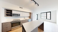 This thoughtfully designed one-bedroom, one-bathroom unit offers a contemporary open-concept layout with clean lines and a neutral palette throughout. The kitchen is a standout feature, equipped with stainless steel appliances including a range, microwave, dishwasher, and a full-size refrigerator. Two-tone cabinetry — white upper cabinets paired with warm wood-grain lower cabinets — lines the wall, complemented by a herringbone tile backsplash and light quartz countertops. A central island with built-in shelving provides additional storage and prep space, seamlessly connecting the kitchen to the open living area. Light hardwood flooring runs throughout, lending warmth and continuity to the space. Track lighting overhead enhances the modern aesthetic. A large window in the living area invites natural light and offers views of the surrounding neighborhood, while a balcony provides an outdoor retreat. The bedroom is a comfortable, well-proportioned room with a black-framed window and hardwood floors, supported by two closets for ample storage. The bathroom features large-format tile flooring, a wood-grain vanity with quartz countertop, an undermount sink, and a full-width illuminated mirror. A hallway connects the rooms efficiently, completing this well-appointed, modern residence.