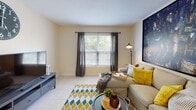 This single-floor residence offers a thoughtfully designed layout that balances comfort and functionality across its key living areas. The living room serves as the heart of the home, featuring light tile flooring, a textured ceiling, and a large window dressed with charcoal curtains that frame views of lush greenery outside. The open, welcoming space provides ample room for seating and entertainment. Adjacent to the living area, the kitchen is a standout feature, showcasing white cabinetry, a decorative tile backsplash, granite countertops, and a central island with bar seating — ideal for casual dining or meal preparation. Black appliances, including a refrigerator, microwave, and range, provide a sleek contrast to the bright cabinetry. The bedroom is a serene retreat, carpeted for warmth and comfort, with a large window dressed in navy curtains that allow natural light to filter in softly. The room offers generous space for a full bedroom suite. The bathroom is clean and functional, featuring a full tub-shower combination with white tile surround, a granite-topped vanity, and tile flooring. Throughout the space, warm neutral wall tones create a cohesive and inviting ambiance.