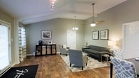 This single-story space presents a thoughtfully designed interior that balances comfort and functionality. The most striking architectural feature is the vaulted ceiling, which adds a sense of openness and airiness to the room. Soft sage-green walls provide a calming, neutral backdrop throughout the space.

The main area serves as a comfortable lounge or waiting room, anchored by a chevron-patterned area rug atop warm hardwood-style flooring. Seating includes a contemporary dark gray sofa, complemented by two upholstered gray accent chairs arranged to encourage conversation or relaxed waiting. A gold-accented side table adds a refined touch to the arrangement.

Lighting is layered effectively, combining a ceiling fan with an integrated light fixture, a decorative chandelier, and track lighting, ensuring the space is well-lit throughout. Framed artwork adorns the walls, contributing to the polished, professional ambiance.

A sleek black credenza along one wall houses a beverage station, suggesting the space is designed to accommodate guests. A private door provides access to an adjoining area, hinting at additional rooms beyond the main lounge.