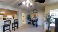 This single-story 2-bedroom, 2-bathroom residence offers a thoughtfully arranged floor plan that maximizes functionality. The main living and dining area features wall-to-wall carpeting and is illuminated by a ceiling fan with integrated lighting, complemented by track lighting that allows for flexible illumination throughout the space. A framed art piece adds a decorative focal point to the dining area, which comfortably accommodates a bistro-style table and chairs.

The kitchen is positioned adjacent to the dining space, separated by a breakfast bar with bar seating — ideal for casual dining or entertaining. The kitchen features wood-toned cabinetry providing ample storage. The layout ensures an open connection between the kitchen and living areas, promoting a cohesive flow throughout the unit.

A hallway visible from the main living area leads to the bathroom and additional rooms, keeping private spaces distinctly separated from the common areas. The textured ceilings are consistent throughout, and neutral wall tones create a clean, versatile backdrop. Overall, the space offers a balanced combination of practicality and comfort suited for everyday living.