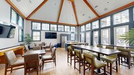 Located at 104 Turnpike Rd in Chelmsford, MA, this expansive community amenity center offers a thoughtfully designed single-floor layout spanning nearly 3,000 square feet of versatile, well-appointed space. The centerpiece is a dramatic clubroom and dining area featuring soaring vaulted ceilings with exposed wood beam accents, shiplap detailing, and floor-to-ceiling windows that flood the space with natural light and provide views of the surrounding community. Bar-height and standard dining tables with contemporary green upholstered seating complement a full kitchen outfitted with stainless steel appliances. A connected lounge area features a stone-accented fireplace, large-format TV, and stylish chartreuse swivel chairs beneath a striking branched chandelier. A game and social room offers shuffleboard, multiple flat-screen TVs, and casual seating beneath matching vaulted ceilings. The dedicated fitness center is comprehensively equipped with treadmills, ellipticals, strength machines, free weights, and benches across hardwood-style flooring, supported by ceiling fans and wall mirrors. A separate yoga and stretching studio provides additional workout space. A private conference room with a wall-mounted display, ergonomic seating, and natural light serves professional needs. The welcoming entry foyer features rich wood floors, stone-tile accent walls, and a mounted display screen.