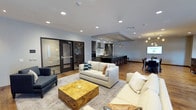 This single-floor space is designed with a contemporary open-concept layout that seamlessly connects multiple functional zones. The living area serves as the central hub, furnished with plush neutral-toned sofas arranged around a distinctive gold-toned wood coffee table, all anchored by a soft area rug. Recessed lighting throughout the ceiling provides consistent, warm illumination across the space.

Adjacent to the lounge area is a fully equipped kitchen featuring dark cabinetry, a large island with bar seating, stainless steel appliances, and a range hood set against a clean tile backsplash. Pendant lighting above the island and bar adds a refined, layered lighting effect.

A dedicated dining area with a dark table and chairs sits nearby, accompanied by additional pendant lighting, making it well-suited for group meals or meetings. Warm hardwood flooring runs throughout, tying each zone together cohesively.

A standout feature is the glass-enclosed private room accessible through dark-framed glass doors, offering a semi-transparent separation ideal for focused work or smaller breakout gatherings. The overall ambiance is modern and polished, balancing comfort with functionality in a well-appointed, multi-use environment.