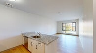 Located at 161 S Huntington Ave in Boston's Jamaica Plain neighborhood, this one-bedroom, one-bathroom residence offers a well-designed layout across a single floor. The open-concept living and kitchen area is anchored by a peninsula with granite countertops and an undermount sink, seamlessly connecting the kitchen to the living space. The kitchen is equipped with stainless steel appliances, including a refrigerator, range, and microwave, complemented by white cabinetry and ample counter space. Light hardwood flooring runs throughout the main living areas, enhancing the clean, modern aesthetic. Large floor-to-ceiling windows flood the living room with natural light while offering views of the surrounding Boston cityscape and treetops. The bedroom features plush carpeting, a dedicated closet, and a window that provides additional natural light and urban views. The bathroom includes a full tub-shower combination finished with classic white subway tile. A separate closet adds practical storage. The overall design is contemporary and neutral, creating a bright, functional living environment well-suited to urban Boston living.