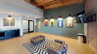 Wag Cafe occupies a single-floor space that masterfully blends industrial heritage with modern, pet-friendly design. The interior is defined by its striking exposed brick walls, soaring ceilings with original heavy timber beams, and visible ductwork that preserves the building's historic character. Large arched windows dressed with blinds line the walls, flooding the space with natural light while adding architectural interest.

The main gathering area features tufted brown leather Chesterfield seating arranged on bold chevron-patterned area rugs, creating an inviting lounge atmosphere. A wall-mounted flat-screen television serves as a focal point in one corner, while colorful, pop-art-style dog portrait paintings adorn the green and white accent walls throughout, reinforcing the café's canine-centric identity.

A dedicated dining area offers tufted upholstered chairs alongside long communal tables, illuminated by a row of distinctive red pendant lights. The café also includes a service counter featuring light granite countertops, dark cabinetry, a stainless steel sink, and reclaimed wood wall art accents. Herringbone-patterned flooring flows consistently throughout, unifying the space's eclectic mix of rustic and contemporary design elements.