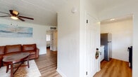 This single-floor unit offers a well-organized layout that makes efficient use of its footprint, as visible in the dollhouse overview. The interior is defined by painted cinder block walls that lend a clean, casual character throughout, complemented by warm wood-look flooring that runs seamlessly across all rooms.

The living area serves as the central gathering space, featuring a large sectional leather sofa in a rich cognac tone, a round wooden coffee table, and a ceiling fan with integrated lighting. A coastal-inspired wall painting adds a relaxed, airy feel to the room.

The kitchenette is compact and functional, outfitted with white cabinetry featuring matte black hardware, a marble-look countertop, a single-basin stainless steel sink, and a full-size stainless steel refrigerator. The white cinder block walls continue here, maintaining a consistent aesthetic.

Two bedrooms provide comfortable accommodations, each furnished with a full-size bed, wooden dresser, and a window with blinds that allow for natural light and privacy. The rooms share the same wood-look flooring and painted block wall construction seen throughout the unit.

The bathroom features white cinder block walls, a towel bar with matte black hardware, and essential fixtures. A central entryway with a classic paneled white door connects the living space to the kitchen, making the overall flow of the unit practical and straightforward.