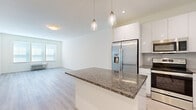 This well-designed 2-bedroom, 1-bathroom residence offers a thoughtfully laid-out single-floor plan with a clean, contemporary aesthetic throughout. The open-concept kitchen and living area forms the heart of the home, anchored by a central island with rich granite countertops and pendant lighting overhead. The kitchen is fully equipped with stainless steel appliances — including a French door refrigerator with ice maker, electric range, built-in microwave, and dishwasher — complemented by sleek white cabinetry that extends to the ceiling and a subtle tile backsplash. Light wood-style flooring runs seamlessly through the kitchen and living area, enhancing the bright, airy feel of the space. The living room benefits from a wide bank of windows that invite natural light, creating a welcoming and open atmosphere. Both bedrooms continue the consistent flooring and neutral color palette, each featuring windows with blinds and individual climate control units. The spacious primary bedroom includes a walk-in closet for ample storage. The bathroom features marble-style tile flooring, a modern vanity with white countertops, a large mirror, and a tiled walk-in shower stall, rounding out this well-appointed residence.