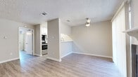This single-floor unit offers a clean, modern aesthetic throughout, anchored by light wood-look flooring and neutral warm-gray walls that create a cohesive, inviting atmosphere. The open-concept living area is a standout feature, centered around a black-tiled fireplace with a white mantel and complemented by a ceiling fan with light fixture overhead. A sliding glass door with vertical blinds floods the space with natural light while providing access to an outdoor area. The living space flows seamlessly into the dining area, which is defined by a half-wall pass-through that connects to the kitchen.

The galley-style kitchen is efficiently designed with white cabinetry, white countertops, and a full suite of stainless steel appliances including a refrigerator, electric range, and dishwasher. A stainless steel sink with a modern pull-down faucet adds a functional touch. The textured popcorn ceilings are consistent throughout the unit, adding a classic residential character. A hallway leads to the bathroom, which features light flooring continuing from the main living areas, and a clean, straightforward layout. Track lighting in one of the rooms provides flexible illumination, rounding out this well-maintained, move-in-ready space.
