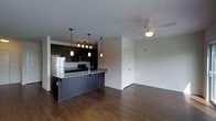 This single-floor space showcases a thoughtfully designed open-concept layout that seamlessly connects the kitchen and living area. The kitchen is a standout feature, outfitted with dark cabinetry, granite countertops, and stainless steel appliances including a refrigerator and microwave. A peninsula with pendant lighting serves as both a functional workspace and a natural divider between the kitchen and the adjoining living space. A track lighting system above the kitchen provides additional illumination, complementing the warm glow of the pendant fixtures. The living area is bright and airy, with crisp white walls and rich, dark hardwood flooring that flows consistently throughout the space, adding warmth and continuity to the design. A ceiling fan with a light kit is centrally installed in the living area, enhancing both comfort and functionality. A closet with double doors is conveniently accessible from the main living space, offering ample storage. The overall ambiance is contemporary and clean, with a neutral color palette accented by the bold contrast of dark cabinetry and flooring, making this a versatile and stylish space.