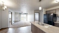 Located at 5910 S Graystone Ave in Sioux Falls, SD, this single-floor, 2-bedroom, 2-bathroom unit offers a practical and well-organized layout. The open-concept living and kitchen area serves as the heart of the space, where wood-look flooring flows throughout. The kitchen features dark wood cabinetry, a dual-basin sink set into a peninsula with pendant lighting above, and black appliances including a refrigerator, range, and over-the-range microwave. The living area extends toward a sliding glass door framed by a ceiling fan, allowing natural light to fill the room and providing access to an outdoor balcony with open views of the surrounding South Dakota landscape. Both bedrooms are carpeted and well-lit by windows with exterior views, each offering a quiet and comfortable retreat with dedicated closet space. The two bathrooms are finished with tile flooring and dark wood vanities, each equipped with a full tub-shower combination. A utility room rounds out the layout, adding functional storage and convenience. The unit's neutral color palette and clean finishes create a cohesive, move-in-ready atmosphere throughout.