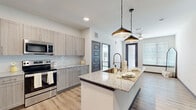 This contemporary single-floor residence is thoughtfully designed with a clean, modern aesthetic throughout. The heart of the space is an open-concept kitchen that flows seamlessly toward the living area. It features a generous central island topped with speckled granite countertops, an integrated undermount sink with a professional-style pull-down faucet, and stylish pendant lights suspended by chain fixtures above. Light wood-toned cabinetry lines the walls, complemented by a white subway tile backsplash and a full suite of stainless steel appliances, including a refrigerator, range, microwave, and dishwasher. Light hardwood-style flooring runs throughout the kitchen and living areas, adding warmth and continuity to the space. In-unit laundry is conveniently accessible near the kitchen. The bedrooms are carpeted for comfort and feature large windows that invite natural light and outdoor views. Charcoal-framed doors provide a modern contrast against the neutral wall tones. The bathroom is a standout feature, offering a wide double-vanity with granite countertops, light wood cabinetry, a large wall mirror with overhead lighting, and direct access to the bedroom, creating a practical and elegant en-suite feel.