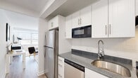This efficiently designed one-bedroom, one-bathroom unit offers a smart and functional layout across a single floor. The open-concept living and sleeping area is bathed in natural light from large windows, creating a bright and airy atmosphere throughout. Light wood-tone flooring runs seamlessly through the main living space, complementing the clean white walls and modern aesthetic.

The kitchen is thoughtfully appointed with crisp white shaker-style cabinetry, granite-look countertops, a classic subway tile backsplash, and stainless steel appliances including a full-size refrigerator, dishwasher, and built-in microwave. A deep stainless steel sink with a flexible-neck faucet adds both style and practicality.

The bedroom area is generously proportioned and defined by an open shelving unit that serves as a stylish room divider, providing storage while maintaining the open feel of the space. A large window floods the room with natural light, enhancing the calm and minimalist ambiance.

The bathroom features light-toned tile flooring, white cabinetry with under-sink storage, a mirrored medicine cabinet, and a shower/tub combination. The all-white palette keeps the space feeling clean and fresh. A connecting hallway ties the rooms together, completing this well-organized and modern unit.