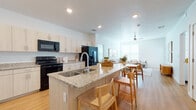 This single-floor residence is designed with a clean, contemporary aesthetic that emphasizes open living and cohesive finishes throughout. The heart of the home is the open-concept kitchen and living area, where a large central island with granite countertops and an integrated sink serves as both a functional workspace and casual dining spot, accommodated by light wood bar stools. The kitchen features light wood cabinetry with brushed metal hardware, matching granite countertops along the perimeter, and black appliances including an electric range, built-in microwave, and refrigerator. Recessed lighting and light wood-style flooring carry through the kitchen and into the living area, which benefits from natural light through large windows and is finished with a ceiling fan overhead.

The bedrooms are carpeted and bathed in natural light, each featuring a ceiling fan, neutral walls, and windows with blinds. Both bathrooms echo the home's consistent design language, showcasing light wood vanity cabinetry, granite countertops, large mirrors, and wood-style flooring. One bathroom includes a shower-tub combination. Throughout, the residence maintains a bright, neutral palette that creates a cohesive and welcoming atmosphere.
