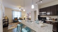 Located in Apalachee, this single-floor residence offers a thoughtfully designed open-concept layout that seamlessly connects the kitchen and living areas. The kitchen is a standout feature, boasting granite countertop surfaces, dark espresso cabinetry, a sleek black range, stainless steel appliances, and a breakfast bar with seating — ideal for casual dining or entertaining. Pendant lighting hangs above the bar, adding warmth and style to the space, while track lighting over the kitchen provides focused task illumination. The living area flows naturally from the kitchen and is bathed in natural light from large windows dressed with elegant draping curtains. A ceiling fan overhead ensures comfort year-round. The space features wood-style flooring throughout, adding a touch of warmth and continuity to the open floor plan. The overall ambiance is modern yet welcoming, with neutral walls that complement the colorful, curated furnishings. A walk-in closet is visible just off the main living area, adding practical storage to the well-organized layout. This residence combines functional design with attractive finishes to create a comfortable and stylish living environment.