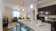 Located in Apalachee, this single-floor residence offers a thoughtfully designed open-concept layout that seamlessly connects the kitchen and living areas. The kitchen is a standout feature, boasting granite countertop surfaces, dark espresso cabinetry, a sleek black range, stainless steel appliances, and a breakfast bar with seating — ideal for casual dining or entertaining. Pendant lighting hangs above the bar, adding warmth and style to the space, while track lighting over the kitchen provides focused task illumination. The living area flows naturally from the kitchen and is bathed in natural light from large windows dressed with elegant draping curtains. A ceiling fan overhead ensures comfort year-round. The space features wood-style flooring throughout, adding a touch of warmth and continuity to the open floor plan. The overall ambiance is modern yet welcoming, with neutral walls that complement the colorful, curated furnishings. A walk-in closet is visible just off the main living area, adding practical storage to the well-organized layout. This residence combines functional design with attractive finishes to create a comfortable and stylish living environment.