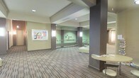 This single-floor space presents a thoughtfully designed interior that blends functionality with aesthetic appeal, characteristic of a professional or institutional setting. The open-plan layout is anchored by sturdy square columns finished in a rich dark tone, which provide structural definition while contributing to the overall design scheme. The flooring features a distinctive brick-patterned tile in muted earthy tones, adding texture and visual interest underfoot. Walls are painted in a soft sage green, creating a calm and welcoming ambiance throughout the space. The ceiling is smooth and well-lit, with recessed lighting ensuring even illumination across the area. A standout feature is the prominently displayed large-scale framed artwork on the wall, adding a creative focal point to the environment. Smaller framed pieces are arranged along the far wall, reinforcing a gallery-like atmosphere. A low, curved railing suggests a change in floor elevation or a mezzanine-style feature nearby. Seating elements and a small table are visible, indicating a lounge or waiting area. The space conveys a professional yet inviting character, well-suited for receiving visitors or facilitating informal gatherings.
