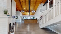 This two-story commercial space makes an immediate impression with its dramatic vaulted ceilings adorned with rich, exposed wood beams and tongue-and-groove planking. The expansive main level centers around a striking reception desk with wood-panel detailing, flanked by statement globe chandeliers that hang from the soaring ceiling above. Large windows flood the space with natural light, while wood-look flooring runs throughout, tying the design together cohesively.

A white-railed staircase leads to the upper level, which features an open loft-style layout complete with a distinctive circular porthole window that adds architectural character. This upper floor also includes a long conference table beneath the vaulted beams, ideal for collaborative gatherings or meetings.

The ground floor offers additional functional areas, including a private meeting room with a round table and street-facing windows, as well as a comfortable lounge area with upholstered seating. A fully equipped galley-style kitchen features white cabinetry, quartz countertops, stainless steel appliances, and subway tile accents, providing a clean and practical workspace amenity.

Throughout the space, coastal-inspired artwork and lush greenery contribute to a bright, welcoming ambiance that balances professional functionality with a relaxed, modern aesthetic.