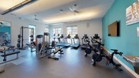 The Ledgewood Fitness Center in Netcong, New Jersey is a thoughtfully designed, single-floor fitness facility that offers a comprehensive workout environment for all fitness levels. The space features a clean, modern aesthetic with crisp white walls accented by bold teal feature walls, durable rubber flooring, and ceiling fans that contribute to a comfortable workout atmosphere.

Large windows line the perimeter of the facility, flooding the space with natural light and offering views of the surrounding community — an especially welcome feature in the cardio zone, where treadmills, ellipticals, and upright bikes are positioned to take full advantage of the outdoor scenery. This area also includes spin bikes for high-intensity cycling workouts.

The strength training section is equally well-appointed, featuring a power rack with barbells and weight plates, flat benches, and a dedicated free weight area with dumbbells neatly racked along mirrored walls that enhance the sense of spaciousness. A multi-station cable machine provides additional options for resistance training.

The facility also includes stability balls and functional training accessories, rounding out the full range of fitness offerings. A glass-panel entry connects to an adjacent corridor, while motivational artwork decorates the teal accent walls, adding an energetic and welcoming character to the space overall.