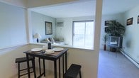 This single-floor residence offers a thoughtfully arranged open-concept layout that connects the key living areas in a natural and functional flow. The dining area features a dark-toned high-top table with bar-style seating, set atop tile flooring with woven placemats and simple place settings that lend a casual yet inviting atmosphere. A half-wall partition defines the space without fully closing it off, creating a sense of openness while maintaining a subtle sense of separation between areas. Beyond the partition, the living room is bathed in natural light streaming through a large sliding glass door that offers views of an outdoor space. The living room features soft, neutral-toned seating and is accented by a wall-mounted air conditioning unit and a framed piece of wall art, contributing to the room's understated charm. The walls throughout are painted in a soft, calming blue-gray tone that enhances the light and airy feel of the interior. Adjacent to the living room, a carpeted area extends the usable living space, decorated with a tall decorative plant in a warm terracotta vase, adding a natural element to the overall aesthetic. The space strikes a balance between simplicity and comfort.