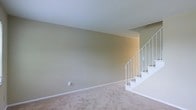 This two-floor space offers a straightforward and functional layout designed for comfortable living. The main level features wall-to-wall carpeting in a warm beige tone, providing a soft and inviting foundation throughout the living area. The walls are finished in a neutral sage-green hue, contributing to a calm and understated ambiance that lends itself well to a variety of interior styles.

A white-railed staircase is prominently positioned along one side of the main living area, connecting the two floors with a clean, classic design. The staircase features white balusters and carpeted treads that complement the flooring below, creating a cohesive visual transition between levels. The upper landing is partially visible from the main floor, hinting at additional living space above.

The main living area is open and unobstructed, offering flexible arrangement possibilities. White baseboards frame the perimeter of the room, adding a crisp contrast to the neutral wall color. The space is well-lit with an even, diffused light that highlights the clean lines and simple finishes throughout. Overall, the space presents a blank canvas well-suited for personalization.