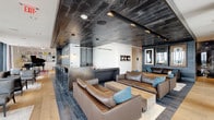 Located at 800 P Street NW in Washington D.C.'s vibrant Logan Circle neighborhood, this upscale amenity lounge occupies an elevated floor with sweeping panoramic views of the city skyline and surrounding treetops. The space is defined by a striking contrast of materials — a richly textured, dark reclaimed wood ceiling anchors the bar and lounge area, while light wide-plank hardwood flooring extends throughout the seating zones, creating a dynamic interplay of warmth and sophistication.

The central bar area features a sleek counter with bar stool seating, framed by exposed brick-style tile and pendant lighting. Adjacent to the bar, deep brown leather sectional sofas with teal accent pillows provide a welcoming gathering space. A grand piano and curated bookshelf set the tone in a secondary lounge area, where clusters of dark leather armchairs are arranged around intimate side tables.

Floor-to-ceiling windows along one full wall flood the space with natural light and offer unobstructed views of Washington D.C. Glass doors open directly onto a furnished outdoor terrace, extending the social experience outside. A corridor lined with glossy dark panels — featuring Marshall branding — leads to well-appointed restrooms with double-sink vanities, large-format tile flooring, and artistic mirrored backdrops. The overall ambiance is modern, curated, and music-inspired.