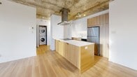Located at 1770 Rue Joseph-Manseau in Montréal, this contemporary 2-bedroom, 1-bathroom unit showcases an industrial-modern aesthetic defined by striking exposed concrete ceilings throughout and warm light hardwood flooring. The open-concept kitchen and living area forms the heart of the unit, where a sleek island with light wood cabinetry, a white countertop, stainless steel appliances, and an overhead range hood anchor the kitchen. The living area enjoys generous natural light through large windows that frame panoramic city skyline views, with access to a private balcony. A hallway connects the main living space to the two well-proportioned bedrooms, each featuring the same concrete ceiling detail, hardwood floors, and windows overlooking the city. Three closets provide ample storage throughout the unit. The clean, white bathroom includes a full bathtub with a glass shower enclosure, a wood-accented vanity, and tile flooring. A dedicated in-unit laundry area with stacked washer and dryer adds everyday convenience. The entry hallway offers a smooth transition into the unit, reinforcing the cohesive, minimalist design that characterizes the entire space.