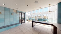 Located at 2439 Beatrice St in Dallas, Texas, this single-floor space showcases a contemporary architectural design that seamlessly blends indoor and outdoor living. The centerpiece of the space is a generously sized semi-covered terrace enclosed by tall, floor-to-ceiling glass panels framed by crisp white structural columns. This open-air area is furnished with light blue upholstered seating, colorful woven accent chairs, and a dark-topped communal table, all bathed in abundant natural sunlight. Brass pendant lights hang overhead, adding a refined, modern touch to the casual outdoor atmosphere. The glass railing system along the terrace perimeter allows for unobstructed panoramic views of the surrounding Dallas cityscape and neighboring greenery.

An interior anteroom or transitional space is distinguished by its bold turquoise large-format tile work covering the walls from floor to ceiling. Built-in concrete benches line the perimeter, and industrial-style bulkhead wall sconces provide warm, utilitarian lighting. Dark-framed glass doors connect this tiled room to the terrace, creating a cohesive flow throughout. Large neutral floor tiles are consistent across both areas, contributing to a clean, unified aesthetic throughout the space.