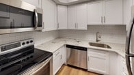 This single-floor unit is designed with a straightforward, functional layout that maximizes the use of available space. The entryway features hardwood-style flooring that transitions into plush wall-to-wall carpeting across the main living area and bedroom. The living space is bright and open, with large windows fitted with horizontal blinds that allow natural light to fill the room while offering privacy control.

The kitchen is a standout feature, showcasing white shaker-style cabinetry, granite countertops, and a classic white subway tile backsplash. Stainless steel appliances—including an electric range, over-the-range microwave, and dishwasher—complement the modern aesthetic. A stainless steel sink with a brushed nickel faucet completes the workspace.

The bedroom is a comfortable, carpeted space with a wide bifold closet offering built-in shelving for organized storage. Neutral gray walls and clean white trim maintain a consistent, understated palette throughout.

The bathroom includes a full-size tub and shower combination with white tile surround and brushed nickel fixtures. Overall, the unit presents a clean, neutral aesthetic with updated finishes and ample natural light.