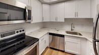 This single-floor unit is designed with a straightforward, functional layout that maximizes the use of available space. The entryway features hardwood-style flooring that transitions into plush wall-to-wall carpeting across the main living area and bedroom. The living space is bright and open, with large windows fitted with horizontal blinds that allow natural light to fill the room while offering privacy control.

The kitchen is a standout feature, showcasing white shaker-style cabinetry, granite countertops, and a classic white subway tile backsplash. Stainless steel appliances—including an electric range, over-the-range microwave, and dishwasher—complement the modern aesthetic. A stainless steel sink with a brushed nickel faucet completes the workspace.

The bedroom is a comfortable, carpeted space with a wide bifold closet offering built-in shelving for organized storage. Neutral gray walls and clean white trim maintain a consistent, understated palette throughout.

The bathroom includes a full-size tub and shower combination with white tile surround and brushed nickel fixtures. Overall, the unit presents a clean, neutral aesthetic with updated finishes and ample natural light.