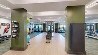 This 1,940-square-foot fitness and wellness facility is thoughtfully designed across a single floor, offering a comprehensive suite of spaces for health and recreation. The main gym area features a combination of light hardwood and plush green carpet flooring, accommodating a wide array of strength and cardio equipment including cable machines, weight benches, treadmills, ellipticals, stationary bikes, and a full dumbbell rack backed by floor-to-ceiling mirrors. Motivational large-scale fitness murals energize the walls, while recessed and linear lighting illuminate the space. A dedicated functional training room provides battle ropes, medicine balls, resistance bands, foam rollers, step platforms, and pegboard-mounted accessories, all set beneath sleek dark linear ceiling lights. An adjacent lounge area offers a welcoming retreat with mid-century modern leather seating, a large wall-mounted display, a fluted green accent wall, and generous natural light from oversized windows facing an outdoor courtyard. The facility is connected by a refined corridor featuring herringbone-patterned hardwood flooring, white paneled millwork, built-in cabinetry, and recessed lighting, creating a polished and cohesive aesthetic throughout the entire space.