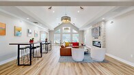 This single-floor community space offers an open-concept layout that seamlessly blends a lounge area, dining zone, and fully equipped kitchen. The centerpiece of the main gathering area is a dramatic vaulted ceiling that rises to a prominent peak, amplifying the sense of spaciousness and volume. A large, multi-panel window at the gable end floods the interior with natural light, complementing the warm light wood flooring found throughout. A floor-to-ceiling stacked-stone fireplace anchors one wall, topped with a flat-screen display, and is flanked by stylish barrel-shade pendant lighting fixtures that add a contemporary touch.

The lounge area features a generous leather sectional sofa, round glass-top coffee table, and modern accent chairs arranged on a patterned area rug. Adjacent to the lounge, tall bistro-style tables with sleek bar stools provide casual seating, while vibrant abstract artwork adorns the neutral gray walls. A glass-paned door leads to a separate, carpeted workspace or office area.

The kitchen is well-appointed with warm maple cabinetry, quartz countertops, stainless steel appliances including a full-size refrigerator and microwave, and a peninsula with leather bar stools — ideal for a communal setting. The cohesive design throughout creates a welcoming, functional, and aesthetically refined environment.
