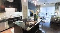 This thoughtfully designed single-floor space makes a strong impression through its open-concept layout and bold interior choices. The kitchen is a standout feature, outfitted with sleek dark cabinetry, dark stone countertops, and a spacious island that serves as both a functional prep area and an entertaining centerpiece. A built-in sink, ample storage, and track lighting overhead contribute to the kitchen's polished, modern aesthetic. The kitchen flows naturally into the dining area, where a wooden dining table and industrial-style seating create an inviting setting for shared meals. A dramatic navy accent wall anchors the living and dining space, adding depth and personality to the interior. Warm hardwood floors run throughout, grounding the darker design elements with natural richness. Pendant lights in a soft rose hue hang above the kitchen island, complementing the ceiling fan that extends comfort into the adjacent living area. Patterned curtains frame large windows and a glass-railed balcony door, allowing natural light to pour in while offering views of the outdoors. The overall ambiance balances contemporary sophistication with a welcoming warmth, making this a well-appointed space suited for both everyday living and effortless entertaining.