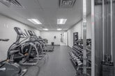 This fitness center at 9001 Patterson Ave in Richmond, Virginia, offers a well-equipped, single-floor exercise facility designed to support a comprehensive workout routine. The space features a drop-tile ceiling with recessed fluorescent lighting that evenly illuminates the room, complemented by neutral-toned walls and durable rubber flooring throughout — ideal for heavy equipment and high-traffic use.

The cardio zone is well-stocked with multiple treadmills and elliptical trainers arranged in a row, allowing users to exercise with ample personal space. A large wall-mounted mirror runs along one side of the room, visually expanding the space and allowing for form checks during workouts. A cluster of spin bikes occupies a dedicated area, providing options for group-style cycling sessions.

The strength training section includes a multi-tiered dumbbell rack stocked with a wide range of free weights, a cable pulley machine suitable for a variety of functional exercises, a weight bench, and suspension training equipment. A wall-mounted sandbag and medicine ball rack rounds out the strength offerings.

A dedicated utility corner provides a dual-basin water fountain and a sanitation station for equipment wipe-downs, ensuring a clean and hygienic environment. Access to adjacent facilities is available through a connected doorway, making this a convenient and fully functional fitness space.
