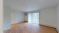 This single-floor unit is designed with a clean, neutral aesthetic throughout. The main living area is generously proportioned, showcasing light-toned hardwood flooring that extends across the space. A large sliding glass door, dressed with vertical blinds, serves as the focal point of the room, drawing in abundant natural light and providing views of the outdoors. Baseboard heating runs along the perimeter walls, and a wall-mounted air conditioning unit ensures year-round climate comfort.

A secondary room, likely used as a bedroom, features two windows fitted with horizontal blinds, offering additional natural light from multiple directions. Baseboard heating is also present here, maintaining the consistent, practical design language found throughout the unit.

The galley-style kitchen is notably upgraded, featuring white shaker cabinetry with brushed nickel hardware, marble-look quartz countertops, a matching backsplash, and a full suite of stainless steel appliances including a range, refrigerator, and dishwasher.

The bathroom continues the refined finish level with a marble-look vanity countertop, a framed mirror, white tile wainscoting, and classic mosaic tile flooring, blending traditional character with modern updates.
