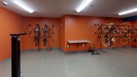 Located at 1989 Cheshire Bridge Rd NE in Atlanta, Georgia, this single-floor bike storage and maintenance room is purpose-built for cyclists. The space is immediately recognizable by its bold burnt-orange accent walls, which contrast with the light gray epoxy flooring and white ceiling, creating a vibrant and functional environment.

The room is equipped with multiple wall-mounted vertical bike racks positioned along the perimeter, allowing for efficient storage of several bicycles without occupying floor space. A freestanding bike repair stand is centrally positioned, providing a convenient station for on-the-spot maintenance. A granite-topped wall-mounted work surface offers additional workspace for tools or accessories.

A set of bright yellow lockers adds a striking visual element near the entrance corridor and provides secure storage for helmets, locks, and other cycling gear. Fluorescent overhead lighting ensures the space is well-lit throughout. The corridor leading into the room features additional wall-mounted hooks and a door providing access to adjacent areas.

Overall, this space is thoughtfully designed to support an active cycling community, combining secure bike storage, maintenance amenities, and personal storage in a compact, clearly organized layout.