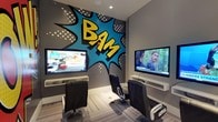 This single-floor entertainment space makes an immediate visual impact with its striking comic book-inspired design. The walls are adorned with large-scale, pop-art murals featuring bold onomatopoeic graphics — including dramatic "POW!" and "BAM!" lettering rendered in vivid yellows, reds, and blues against a dark grey background with classic halftone dot detailing. The design creates a dynamic, energetic atmosphere that sets the tone for the entire space.

The room is equipped with multiple wall-mounted flat-screen televisions positioned at comfortable viewing angles, each supported by sleek white floating shelves. Ergonomic, high-back gaming and lounge chairs are arranged to face the screens, providing dedicated seating for media consumption or gaming sessions. The seating is upholstered in black leather-style material, offering both comfort and a clean aesthetic that complements the bold surroundings.

Recessed ceiling lighting provides even illumination throughout the space, while the striped, multi-toned carpet flooring adds a subtle textural contrast to the room's vivid walls. The overall ambiance is lively and immersive, making this space well-suited as a communal gaming lounge, media room, or entertainment hub designed to energize and engage its users.