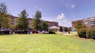 Located at 1001 Hayes Ln in Holly Springs, North Carolina, this vibrant outdoor community space is part of a well-designed mixed-use development that blends classic architectural style with inviting public amenities. The multi-story brick buildings that frame the area feature green awnings and traditional detailing, lending the environment a charming, town-center character that is both welcoming and functional.

At the heart of the space stands a covered gazebo with a pitched roof, serving as a focal point for the surrounding open lawn. The expansive, well-maintained green turf provides ample room for outdoor gatherings and leisurely activities, bordered by mature trees that offer natural shade and visual depth against the open Carolina sky.

Complementing the central green, a carefully landscaped garden area features raised wooden planter beds, mulched pathways, and dense ornamental shrubs that create a sense of enclosure and tranquility. The layered plantings—including tall evergreens, colorful deciduous shrubs, and low-growing groundcover—add rich texture and seasonal interest to the grounds.

Parking is accessible along the perimeter, making the space convenient for visitors. The overall layout reflects thoughtful community planning, balancing retail and residential uses with generous outdoor areas designed for public enjoyment. This space presents a cohesive, pedestrian-friendly environment ideal for community engagement in Holly Springs.