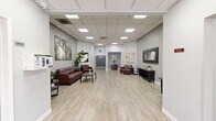 Located at 3560 Flora Vista Ave in Santa Clara, CA, this single-floor commercial suite offers a thoughtfully designed open-plan layout that balances professional functionality with a warm, welcoming ambiance. Light wood-style luxury vinyl flooring runs throughout the space, complementing the soft gray walls and creating a clean, modern aesthetic.

The main area serves as a generously sized lounge and reception zone, furnished with rich burgundy leather seating, dark wood accent tables, and a large rectangular coffee table. A full-length framed mirror anchors one wall, while an open storage console beneath it adds both practicality and style. Lush potted greenery and fresh floral arrangements are thoughtfully placed throughout, adding life and color to the neutral palette.

Large-scale canvas artwork adorns the walls, including nature-inspired prints and cityscapes, contributing to a curated, gallery-like feel. A drop ceiling with recessed skylight panels diffuses bright, even lighting across the entire floor.

Toward the rear of the suite, a corridor leads to additional private areas, including what appears to be a treatment or service room, as well as a niche alcove housing a dark wood cabinet with a stone countertop — ideal for reception or check-in use. The glass entry doors at the front of the suite allow natural light to filter in, enhancing the open and airy character of this well-appointed commercial space.