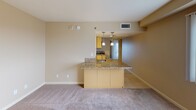 This single-floor unit at 801 Actionable City Blvd, #716 in National City offers a straightforward, functional layout with consistent warm neutral tones throughout. The open living area features plush wall-to-wall carpeting and receives generous natural light, particularly notable near the entry where sunlight casts bright patterns across the floor.

The kitchen is defined by a wraparound peninsula topped with speckled granite countertops, complemented by matching cabinetry in a warm yellow-toned finish. Pendant lighting hangs above the peninsula, adding a focused decorative element to the space. The kitchen flows openly into the living area, enhancing the sense of connectivity between spaces.

A separate carpeted room features a full-width sliding glass door fitted with vertical blinds, allowing filtered natural light and providing access to an outdoor view. This versatile space could serve as a bedroom or secondary living area.

The bathroom is finished with tile flooring, a granite-topped vanity with matching cabinetry, a large mirror, and a glass-enclosed walk-in shower with tiled surround. The unit is currently unfurnished, offering a clean slate for personalization.
