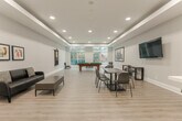 Located at 65 Lillian St in Toronto's midtown neighbourhood, this amenity space serves as a well-appointed communal lounge designed for both recreation and relaxed gathering. The open-concept layout is finished with light wood-look flooring throughout, complemented by soft grey walls and recessed tray ceilings that lend a clean, contemporary feel.

The main area is anchored by a full-size billiards table positioned near expansive floor-to-ceiling windows that invite generous natural light and offer views of the surrounding greenery. A wall-mounted flat-screen television is paired with a long communal dining table and modern chairs, creating a versatile setting for group activities or casual meetings. A dedicated lounge corner features comfortable black upholstered seating arranged around a central coffee table, ideal for relaxed conversation.

Adjacent to the main lounge, a separate seating area provides additional space for quieter gatherings, accented by framed artwork and recessed lighting. The galley-style kitchen is equipped with stainless steel appliances including a full-size refrigerator and stove, white cabinetry with clean-lined hardware, a white countertop, and tile flooring — offering full functionality for catering or resident use. Together, these spaces form a thoughtfully designed amenity suited for a variety of social occasions.