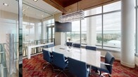 Located at 710 12th Street in Arlington, Virginia, this sophisticated commercial office suite offers a refined, high-end environment designed for professional use. The space is defined by a seamless blend of warm and contemporary materials, including rich wood-paneled ceilings that contrast elegantly with crisp white floors and light stone surfaces.

A standout feature is the formal conference room, enclosed by full-height glass walls that maintain visual openness while providing privacy. The room is anchored by a substantial white stone conference table surrounded by navy blue executive chairs, topped by a striking rectangular crystal pendant chandelier that adds a dramatic focal point. Floor-to-ceiling windows wrap the perimeter, flooding the space with natural light and offering sweeping panoramic views of the Arlington and greater Washington, D.C. skyline.

The open work area features clean-lined marble-top desks equipped with workstations, set against a backdrop of neutral tones and recessed lighting. A lounge area with vibrant orange accent seating adds a pop of color and creates an inviting collaborative zone visible through the glass partitions.

Bold, patterned red carpeting in the conference and lounge areas provides a vivid design counterpoint to the otherwise refined palette. The overall ambiance is polished and executive in character, combining functional workspace with elevated architectural detailing throughout.