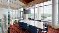 Located at 710 12th Street in Arlington, Virginia, this sophisticated commercial office suite offers a refined, high-end environment designed for professional use. The space is defined by a seamless blend of warm and contemporary materials, including rich wood-paneled ceilings that contrast elegantly with crisp white floors and light stone surfaces.

A standout feature is the formal conference room, enclosed by full-height glass walls that maintain visual openness while providing privacy. The room is anchored by a substantial white stone conference table surrounded by navy blue executive chairs, topped by a striking rectangular crystal pendant chandelier that adds a dramatic focal point. Floor-to-ceiling windows wrap the perimeter, flooding the space with natural light and offering sweeping panoramic views of the Arlington and greater Washington, D.C. skyline.

The open work area features clean-lined marble-top desks equipped with workstations, set against a backdrop of neutral tones and recessed lighting. A lounge area with vibrant orange accent seating adds a pop of color and creates an inviting collaborative zone visible through the glass partitions.

Bold, patterned red carpeting in the conference and lounge areas provides a vivid design counterpoint to the otherwise refined palette. The overall ambiance is polished and executive in character, combining functional workspace with elevated architectural detailing throughout.