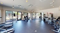 This fitness center at 8100 U.S. 98 in Pensacola, Florida, offers a dedicated exercise room designed to support a comprehensive workout experience. The space is thoughtfully laid out with a durable rubber flooring surface that covers the entire workout area, providing both comfort and safety during exercise.

The room is generously outfitted with cardio equipment, including multiple treadmills and an elliptical machine, alongside a well-rounded selection of strength training stations and cable machines, as well as a stationary bike. Ceiling fans and overhead HVAC units ensure consistent airflow and climate control throughout the space.

Large windows line the perimeter walls, flooding the interior with natural light and offering views of the surrounding outdoor area, including what appears to be a pool area with mature trees. Glass-paneled doors provide additional access points and contribute to the bright, open atmosphere of the room.

A small entryway area serves as a transition into the fitness floor, featuring a wall-mounted TV, a glass-topped table, and vending machines for added convenience. Recessed lighting and ceiling-mounted fixtures supplement the natural light, keeping the space well-illuminated at all hours. The clean, neutral color palette of white walls and light ceilings reinforces a focused and energizing environment suited for fitness and wellness activities.